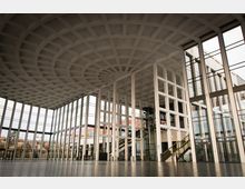 Großer, moderner Innenraum mit hohen Glaswänden und einem symmetrischen, gitterförmigen Deckendesign aus Beton. Im Raum befinden sich ein offenes Treppenhaus, verglaste Fahrstühle und große Fenster mit Blick nach draußen.