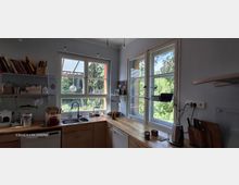 Helle Wohnküche mit hellblauen Wänden, Holzarbeitsplatten und Doppelspüle unter großen Sprossenfenstern. Offene Wandregale mit Gläsern und Vorratsdosen sowie Küchengeräte auf der Arbeitsfläche; durch die Fenster Blick ins grüne Gartenumfeld.