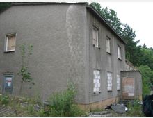 Ein verlassenes, zweistöckiges Gebäude mit grauer Putzfassade; mehrere Fenster sind mit Holz oder Mauerwerk verschlossen. Die Umgebung ist überwuchert mit Pflanzen, und im Hintergrund sind Bäume sichtbar.