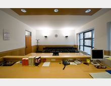 Ein Besprechungsraum mit Schreibtisch im Vordergrund, auf dem Bücher, ein Telefon, ein Bildschirm und Büromaterialien liegen. Im Hintergrund befinden sich Stuhlreihen, eine Garderobe, eine Wanduhr und große Fenster auf der rechten Seite, die Tageslicht hereinlassen.