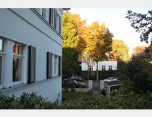 Straßenszene in einer ruhigen Wohngegend mit einem weiß gestrichenen Gebäude im Vordergrund und einem Garten mit Sträuchern. Im Hintergrund stehen Bäume mit herbstlich verfärbtem Laub, dahinter sind weitere weiße Häuser und geparkte Autos zu sehen.