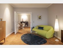 Wohnzimmer in modernem Design mit hellgrauen Wänden und Parkettboden. Im Vordergrund ein hellgrünes Sofa, ein gläserner Couchtisch und ein dunkelgrauer Teppich; im Hintergrund ein Essbereich mit weißen Stühlen und einem Regal, getrennt durch Glastüren.