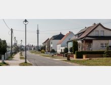 Straßenszene in einem Wohngebiet mit Einfamilienhäusern, die nebeneinander auf beiden Seiten der Straße stehen. Im Vordergrund sind Gehwege, Laternen und gepflegte Vorgärten zu sehen, im Hintergrund ein hoher Funkmast.