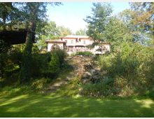 Zweigeschossiges Haus im mediterranen Stil mit heller Fassade, umgeben von dicht bewachsenem Garten und hohen Bäumen; im Vordergrund befindet sich ein Gartenbereich mit einem steinigen Hang und einer angedeuteten Treppe.