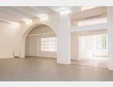 Heller, leerer Innenraum mit weißen Wänden, hohen Decken und einem großen Rundbogen, der zwei Bereiche verbindet. Große Sprossenfenster lassen viel Tageslicht auf den grauen Betonboden; an der Decke sind lineare Leuchten montiert.