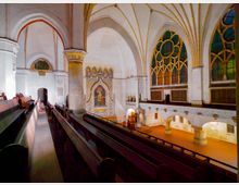 Innenraum einer historischen Kirche mit weißen, gotischen Gewölbedecken und verzierten Spitzbogenfenstern mit farbigem Glas. Holzbänke führen zu einem kunstvoll gestalteten Altar mit einer Ikone, flankiert von massiven Säulen und hohen Wänden.