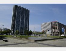 Mehrgeschossige Gebäude in einer urbanen Umgebung, darunter ein hohes Gebäude mit der Aufschrift „Technische Universität“. Im Vordergrund befindet sich ein gepflasterter Platz mit einem rechteckigen Brunnen und mehreren kleinen Wasserspielen, umgeben von Flaggenmasten und Bäumen.