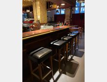 Eine Bar mit einer langen, dunkelroten Theke, auf der Getränke und Gläser stehen. Im Vordergrund befinden sich hohe Hocker mit schwarzen Sitzflächen, im Hintergrund Regale mit Flaschen, eine rot-schwarze Kaffeemaschine und eine Wand mit quadratischen Kacheln.