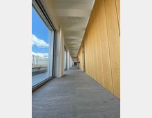 Langer, moderner Flur mit grauem Steinboden und einer Wand aus hellen Holzpaneelen. Links befinden sich bodentiefe Fenster mit Blick auf eine Terrasse und blauen Himmel mit Wolken.