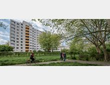 Wohngebiet mit mehrstöckigem Wohnblock im Hintergrund, umgeben von Bäumen und Grünflächen. Im Vordergrund eine Person auf einem Fahrrad sowie zwei Fußgänger auf einem Gehweg.