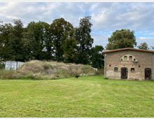 Backsteingebäude mit Holztüren und kleinen Fenstern auf einer Wiese, daneben eine bewachsene Erdhügelstruktur und im Hintergrund Bäume sowie ein Metallcontainer.