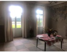 Ein rustikaler Raum mit heller, abgenutzter Wandgestaltung und zwei großen Fenstertüren mit hellen Vorhängen, die nach außen auf eine grüne Landschaft blicken. Im Vordergrund steht ein Tisch mit einer rot-weiß karierten Tischdecke, darauf eine Vase mit Blumen und weitere Gegenstände.