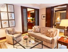 Helles Wohnzimmer mit eleganter Einrichtung, darunter ein gepolstertes Sofa, ein Sessel mit Muster, ein Glastisch mit Blumenarrangement und eine Tischlampe. Im Hintergrund führen dunkle Holztüren zu einem Schlafzimmer und einem Bereich mit Sideboard und Dekorationen.