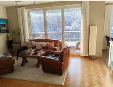 Wohnzimmer mit großen bodentiefen Fenstern, die auf einen verschneiten Balkon führen. Die Einrichtung umfasst braune Ledersofas, einen rustikalen Holztisch auf einem hellen Teppich und eine Stehlampe, mit Parkettboden und weiß-beigen Wänden.