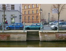 Straßenszene mit parkenden Autos und einem Fußweg neben einer kleinen Wasseranlage mit Geländer und Treppe. Im Hintergrund stehen ein modernes, graues Gebäude und ein rot-gelbes Backsteinhaus mit verzierter Fassade, daneben steht ein Baum ohne Laub.