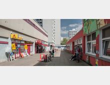 Städtischer Gehweg zwischen zwei Gebäuden, links ein graues Gebäude mit Geschäften wie einem Lottoladen, rechts ein rotes Gebäude mit bemalter Fassade und Fenstern. Im Hintergrund sind mehrere Hochhäuser und ein blauer Himmel sichtbar.