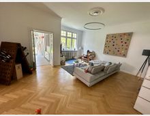 Wohnzimmer mit hellem Fischgrätenparkett, einer großen grauen Sofalandschaft und einem bunten Wandbild. Im Hintergrund befindet sich eine Tür, die zu einem Bereich mit Fliesenboden und großen Fenstern führt.