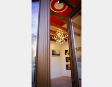 Ein Blick durch eine geöffnete Holztür in einen Ausstellungsraum mit weißen Wänden, Holzdeckenbalken und rotem Deckenbereich mit Stuckornament. Im Raum hängen mehrere Kunstwerke an der Wand, und eine auffällige Lampe mit bunten Lichtern hängt von der Decke.