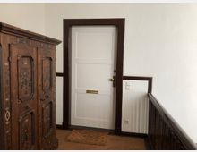 Ein Flur mit weiß gestrichenen Wänden und einer Holztür mit dunkelbraunem Rahmen. Links steht ein großer, reich verzierter Holzschrank, und rechts ist ein Holzgeländer zu sehen. Vor der Tür liegt eine kleine Fußmatte.