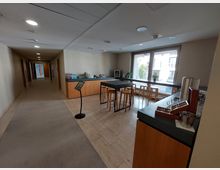 Ein moderner Innenraum mit einem Pausenbereich, bestehend aus einem langen Tresen mit Barhockern, einer Teestation und einem Wasserspender. Große Fenster lassen Tageslicht herein, während ein angrenzender Flur mit Teppichboden vom Bereich wegführt.