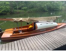 Holzsegelboot mit beige eingerolltem Segel liegt an einem Holzsteg auf einem ruhigen, grünlichen Fluss. Im Hintergrund säumen dicht bewachsene Ufer mit Bäumen und Sträuchern das Gewässer.
