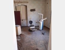 Ein verlassener Raum mit einer alten, ungenutzten Zahnarztpraxis-Ausstattung, bestehend aus einem abgenutzten Behandlungsstuhl und einer Behandlungseinheit. Der Raum hat schmutzige Wände, teilweise verfallene Fensterläden und einen beschädigten Boden.