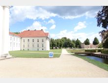 Heller Schlosspark mit breiten Kieswegen, einem kleinen Wasserkanal und gepflegten Grünflächen. Im Hintergrund stehen historische Gebäude mit roten Dächern und heller Fassade, umgeben von Bäumen.