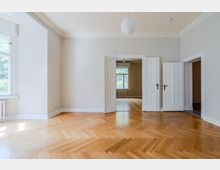 Heller, leerer Altbau-Wohnraum mit hohen Decken, Stuckleisten und Fischgrätparkett. Links ein großes Fenster, mittig eine doppelflügelige Kassettentür zu einem weiteren Raum mit Fenster und Heizkörper.