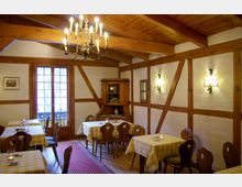 Gemütliches, rustikales Gastrauminterieur mit weiß verputzten Wänden und sichtbaren Holzbalken. Der Raum ist mit Holzmöbeln und gelb karierten Tischdecken ausgestattet, einem Kronleuchter an der Decke und Wandlampen, die warmes Licht spenden; im Hintergrund eine Glastür mit Fenster und ein Holzschrank.