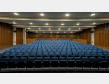 Ein Hörsaal oder Auditorium mit mehreren Reihen blauer Sitze, einer Bühne und einem Rednerpult im Hintergrund. Die Wände sind mit vertikalen Holzpaneelen verkleidet und die Decke verfügt über eingelassene Deckenleuchten.
