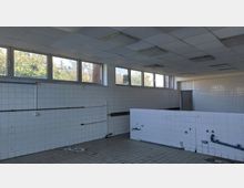 Ein leerer, kachelverkleideter Raum mit weißen Fliesen an den Wänden und am halben Trennelement in der Mitte. Oberhalb befindet sich eine Fensterreihe mit Blick auf Bäume, und an der Decke sind rechteckige Beleuchtungskörper angebracht.