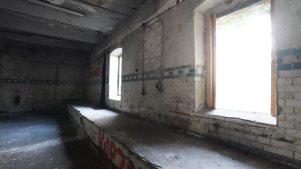 Verlassener Industrie- oder Lagerraum mit stark abgeplatztem Putz, bröckelnder Decke und einer halbhohen gefliesten Wand mit grünem Fliesenband. Zwei hohe Fensteröffnungen lassen grelles Tageslicht herein; entlang der Wand verläuft eine lange, erhöhte Betonplattform mit Graffiti.