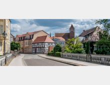 Straßenszene in einer historischen Altstadt mit Fachwerkhäusern, roten Ziegeldächern und Laternen. Im Hintergrund ist eine große Backsteinkirche mit Turm zu sehen, umgeben von Bäumen und grünen Fassaden.