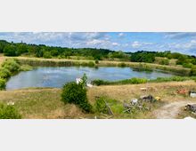 Eine natürliche Landschaft mit einem Teich in der Mitte, umgeben von Wiesen und dichter Vegetation. Im Vordergrund stehen vereinzelt Metallgerüste und Baumaterialien, im Hintergrund erstreckt sich hügeliges Gelände mit Bäumen und vereinzelten Gebäuden.