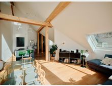 Moderner Wohnraum mit offenem Grundriss in einer Dachgeschosswohnung. Holzbalken, ein Esstisch mit Stühlen, ein kleiner Küchenbereich, ein Sofa, ein Dachfenster und ein Sideboard mit Pflanzen und Schallplatten sind sichtbar.