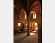 Gewölbter Innenraum aus rotem Ziegelstein mit gotischen Bögen und einer zentralen Säule. Im Raum stehen Holzbänke und Tische auf einem gefliesten Boden, zusammen mit kleinen, bogenförmigen Fenstern, die gedämpftes Licht hereinlassen.