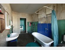 Altes Badezimmer mit freistehender Badewanne und Duscharmatur vor einer Wand aus blauen Fliesen sowie einem Waschbecken mit Spiegel links. Abgenutzte, fleckige Wände in Grün und Beige, dunkler Holzboden und eine offene Tür führen in einen angrenzenden Raum.