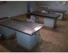 Raum mit gekacheltem Boden und Wänden, ausgestattet mit mehreren Metalltischen auf gefliesten Sockeln; im Hintergrund sind zwei Waschbecken an der Wand montiert.