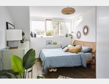 Helles Schlafzimmer mit einem Bett mit geflochtenem Holz-Kopfteil, gedeckt mit blauer Bettwäsche und mehreren Kissen. Der Raum hat weiße Wände, Holzboden, große Fenster mit Blick ins Freie und eine Kommode mit Dekoration sowie Pflanzen.