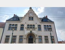 Fassade eines historischen Gebäudes mit hellem Putz, verzierten Fenstern und einem zentralen Giebel, der eine Uhr enthält. Vor dem Eingang mit Rundbogen stehen zwei Laternen, und das Dach zeigt Gauben mit dunklen Ziegeln.