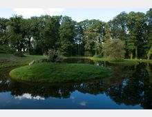 Ein kleiner, von Bäumen umgebener Teich in einer parkähnlichen Landschaft, mit einer grasbewachsenen Insel in der Mitte und Spiegelungen der Bäume auf der Wasseroberfläche. Der Himmel ist teilweise bewölkt, und die Umgebung wirkt ruhig und naturbelassen.