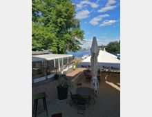 Außenbereich eines Restaurants mit runden Tischen und Stühlen auf Kiesboden, überdacht von Sonnenschirmen mit der Aufschrift „Maisel's Weisse“. Im Hintergrund ein großer Baum, der Blick öffnet sich auf einen See oder Fluss mit Booten und bewaldetem Ufer, unter einem Himmel mit vereinzelten Wolken.