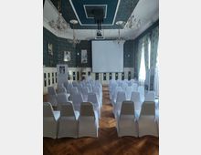 Ein Veranstaltungsraum mit Holzfußboden, einer großen Leinwand an der Stirnwand und mehreren Reihen weiß eingedeckter Stühle. Der Raum ist mit Kronleuchtern, dekorativen Wänden und Vorhängen gestaltet; an den Wänden hängen Bilder.