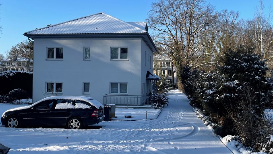 Verschneiter Innenhof mit breiter Zufahrt und Reifenspuren, links ein zweigeschossiges weißes Wohnhaus mit Walmdach und mehreren Fenstern. Rechts säumen Hecken und kahle Bäume den Weg; im Hintergrund weitere Wohngebäude unter blauem Himmel.