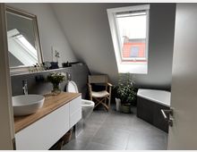 Modernes Badezimmer unter einer Dachschräge mit einer großen Dachfensteröffnung. Zu sehen sind eine Waschtischkombination mit Holzplatte, ein wandhängendes WC, eine Eckbadewanne, ein Stuhl, Topfpflanzen und ein großer Spiegel.
