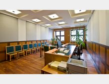 Moderner Büroraum mit Holzboden, hölzernen Wandverkleidungen und eingelassenen Deckenleuchten. Der Raum enthält eine Reihe von Stühlen mit grün gepolsterten Sitzflächen an einer Wand, einen großen Schreibtisch mit Computer-Arbeitsplätzen sowie mehrere Zimmerpflanzen.