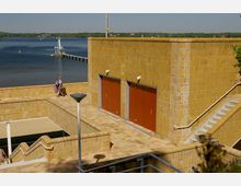 Gelbes Gebäude mit Fliesenfassade und zwei roten Türen, umgeben von Treppen und Geländern, auf einer erhöhten Plattform am Wasser. Im Hintergrund ein See mit einem Bootsanleger und bewaldeter Uferlandschaft.