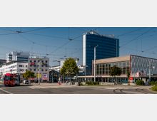 Straßenszene in der Innenstadt von Cottbus mit der markanten "Stadthalle Cottbus" rechts im Bild. Im Hintergrund mehrere moderne Büro- und Geschäftsgebäude, dazwischen eine Straßenbahnhaltestelle mit einer roten Straßenbahn im Vordergrund, umgeben von Fußgängern und Bäumen.