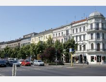 Straßenszene mit Altbauten im Gründerzeitstil, darunter ein weißes Gebäude mit Kuppeldach, sowie weiteren farbigen Gebäuden und Baumbepflanzung entlang der Straße. Im Vordergrund eine Straße mit wartenden Autos und eine U-Bahn-Markierung mit einem „U“-Schild.