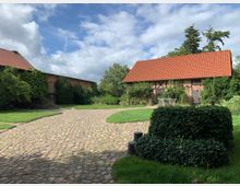 Ein gepflasterter Hof mit zwei rustikalen Backsteingebäuden mit roten Ziegeldächern, umgeben von Grünflächen, Bäumen und Kletterpflanzen. Links und rechts des Bildes sind die historischen Gebäude sichtbar, dazwischen erstreckt sich ein großer, offener Raum.